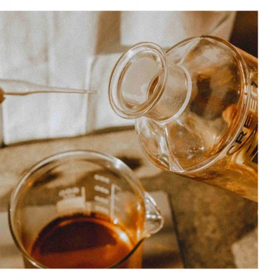 A glass bottle is pouring liquid into a beaker, with a pipette visible on the left and the word CREED printed on the bottle.
