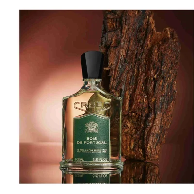 A bottle of Creed perfume is shown next to a piece of bark, with the label reading CREED BOIS DU PORTUGAL DE PERE EN FILS DEPUIS 1760 FROM FATHER TO SON SINCE 1760 100ML 3.33 FL. OZ.