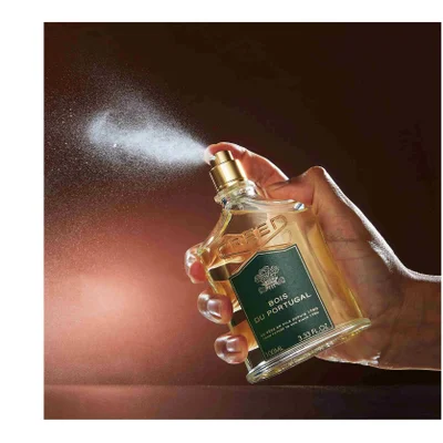 Hand spraying a Creed Bois du Portugal fragrance bottle with visible spray mist and text that reads CREED BOIS DU PORTUGAL DE PERE EN FILS DEPUIS 1760 FROM FATHER TO SON SINCE 1760 100ML 3.33 FL. OZ.