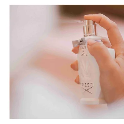 A hand sprays a white perfume bottle with mist coming out showing the text LOVE IN WHITE and CREED on the bottle.