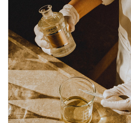 Person in white gloves holds a Creed bottle labeled CREED EAU DE PARFUM IMPERIAL above a beaker with a dropper.