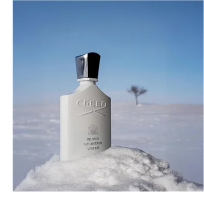 A white fragrance bottle with a silver cap stands against a black background, with a glowing green lime slice behind it and water streams flowing around them, with the text CREED, a crest, and SILVER MOUNTAIN WATER visible on the bottle.
