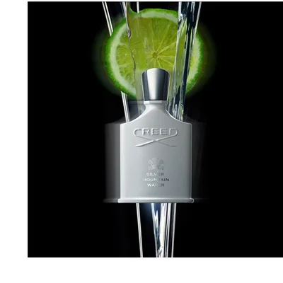 A white fragrance bottle with a silver cap stands against a black background, with a glowing green lime slice behind it and water streams flowing around them, with the text CREED, a crest, and SILVER MOUNTAIN WATER visible on the bottle.