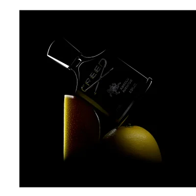 A dark bottle of Creed Absolu Aventus perfume with a rider on a horse logo, marked 75 ML 2.5FL.OZ., is tilted over two citrus fruits.