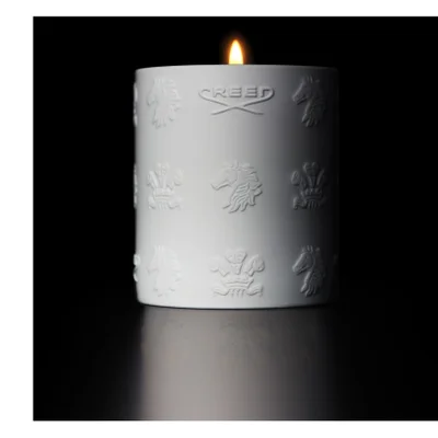 A lit white candle with embossed designs and the text CREED sits on a reflective surface in a dark setting.