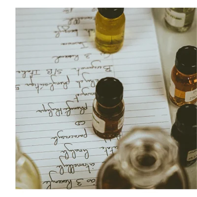 Small glass bottles filled with liquids rest on a lined paper containing handwritten notes for perfume formulation including Jasmine, 3 Risorol, Bergamote S/B Italie, Benzyl phenylacétate 5, Muguet, Phenyl Ethylique, CD, Paracrésyl, Linalyl, Citronellol, ao 3 Hexenyl, and labels on bottles displaying Essence, 24, 2140.