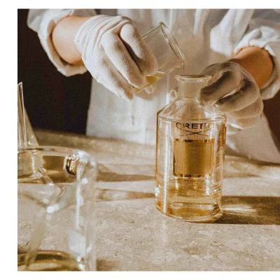 Person wearing gloves pours liquid from a beaker into a glass bottle with the label CREED Millesime Imperial 1760.