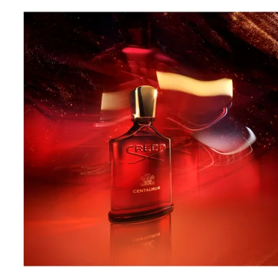A red perfume bottle with a gold cap is shown against a fiery orange background with gold sparkles, with the text CREED DELPHINUS visible on the bottle.