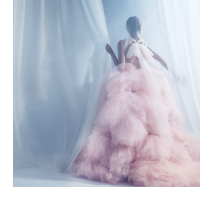 A woman in a voluminous pink dress stands behind a sheer white curtain.