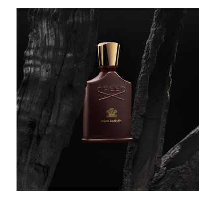A dark brown perfume bottle with a gold cap and embossed gold text CREED and OUD ZARIAN is centered on a dark background between two dark, rough charred wood pieces.
