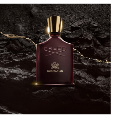 A dark red perfume bottle with a gold cap is illuminated on a textured black surface, with golden liquid flowing around it, featuring the embossed text CREED and OUD ZARIAN.