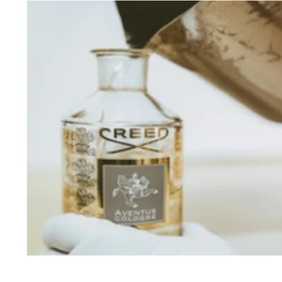 A hand in a white glove holds a clear perfume bottle labeled Creed Aventus Cologne, as liquid is poured into it.