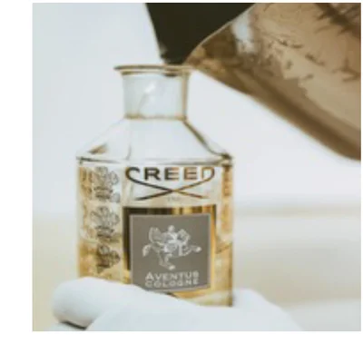 A gloved hand holds a glass bottle of Creed Aventus Cologne as liquid is poured into it from a jug.