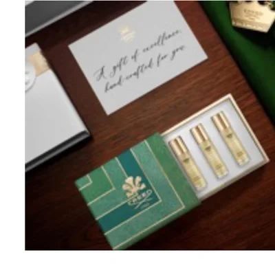 A green and gold patterned gift box, open to show three small gold-topped fragrance bottles, sits on a wooden surface next to a white card and a white gift box; the card reads A gift of excellence, hand-crafted for you, and the green box features a floral emblem and the word CREED.