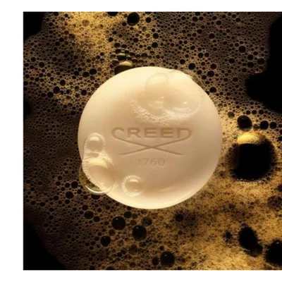 A round, light-colored bar of soap with some bubbles on its surface rests in dark golden-brown foam, featuring the embossed text CREED with a logo and 1760, against a black background.