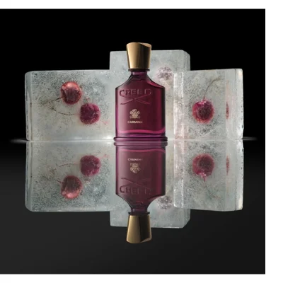 A dark red perfume bottle with a gold cap stands centered between ice blocks containing frozen red cherries, with its reflection visible below, featuring the text CREED and CARMINA.