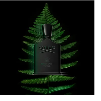 A black Creed perfume bottle with a dark cap rests on dark green fern leaves against a black background, with visible text Creed and Green Irish Tweed on the bottle.