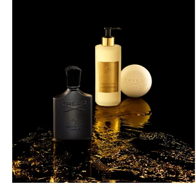 A black Creed perfume bottle, a cream-colored lotion bottle with a gold pump, and a round cream-colored bar of soap from the Green Irish Tweed line are displayed on a reflective black surface with shimmering gold liquid; the perfume bottle reads CREED, GREEN IRISH, TWEED, the lotion bottle reads CREED, 1760, GREEN IRISH TWEED, HAND & BODY WASH, SAVON LIQUIDE MAINS ET CORPS, and the soap reads CREED, 1760.