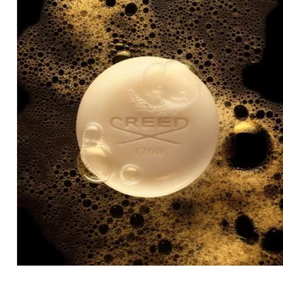 A round bar of light-colored soap with small clear bubbles on its surface, surrounded by rich golden-brown foam, displays the text CREED 1760.