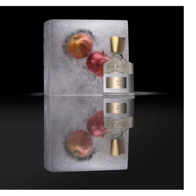 A clear perfume bottle with a gold cap and gold CREED text on the neck stands next to a block of ice containing two red and yellow apples, reflected on a dark surface, with the bottle label displaying AVENTUS FOR HER EAU DE PARFUM and a figure on horseback.