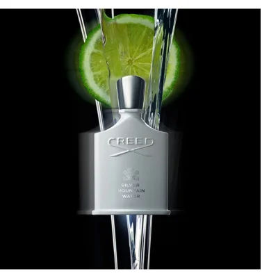 A white Creed Silver Mountain Water perfume bottle with a silver cap and crest logo stands against a black background, with a bright green lime slice and clear water flowing around it, displaying the text Creed, Silver Mountain Water.