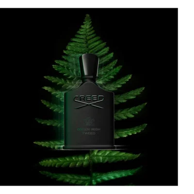 A black fragrance bottle with a black cap stands against a large green fern leaf on a dark background, featuring the text CREED, an emblem, GREEN IRISH, and TWEED.