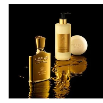 A golden Creed Millesime Imperial perfume bottle stands next to a cream lotion bottle with a black pump and a round cream Creed soap bar, all resting on a reflective surface with golden liquid splashes.