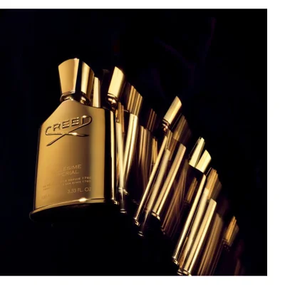 A golden Creed perfume bottle with a reflective cap and engraved text is displayed against a dark background, showing a motion blur effect creating multiple instances of the bottle extending to the right, featuring the text CREED MILLÉSIME IMPERIAL DE PÈRE EN FILS DEPUIS 1760 FROM FATHER TO SON SINCE 1760 100 ML 3.33 FL. OZ.