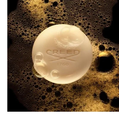 A creamy white round bar of soap with bubbles on its surface and embossed text CREED 1760, surrounded by golden-brown foam on a dark background.