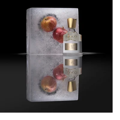 A perfume bottle with a gold cap and a white label featuring the words CREED and AVENTUS stands beside a block of ice encasing two red apples, all reflected on a dark surface.