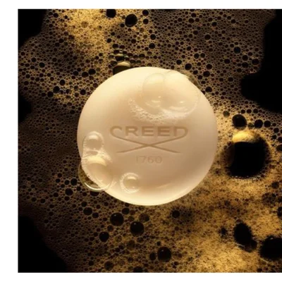 A round, light-colored bar of soap rests in golden-brown foam with many bubbles on a dark background, featuring the embossed text CREED 1760.