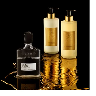 A dark red perfume bottle with a gold cap is illuminated on a textured black surface, with golden liquid flowing around it, featuring the embossed text CREED and OUD ZARIAN.