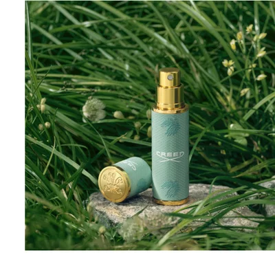 Creed green travel spray with gold cap on stone among grass and small white flowers.